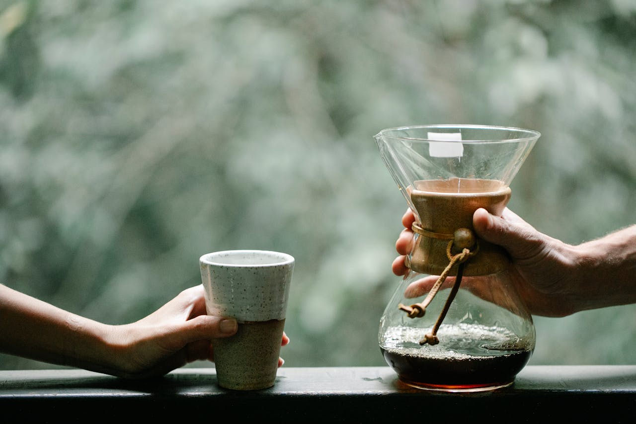 Crop unrecognizable girlfriend and boyfriend with hot drink in glass and Chemex coffee maker on terrace in countryside